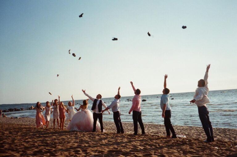A group celebrates a wedding on a beach by joyously throwing shoes in the air, set against a serene ocean backdrop.