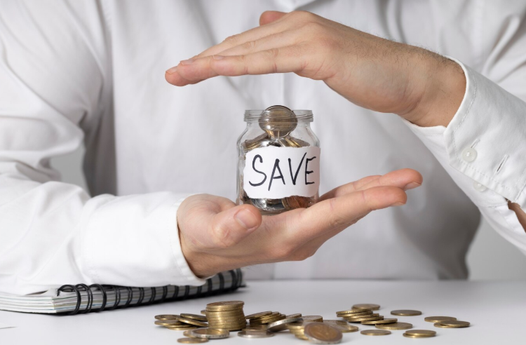 Person holding a coin jar labeled "save" with stacked coins, symbolizing simple swaps to save money each month.
