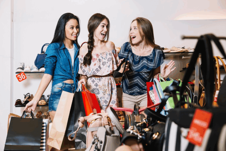 Three young women excitedly shopping in a store clearance section with discounted handbags and shoes